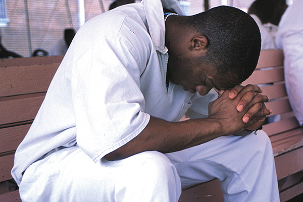 Your gift to provide Bibles can help prisoners like this man sitting on a bench with his head bowed and hands clasped together